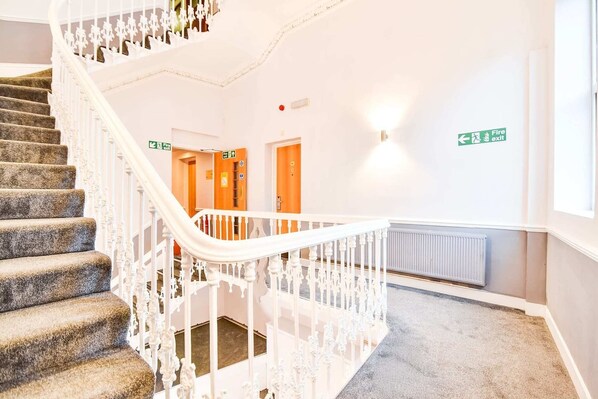 A staircase with a white railing, carpeted steps, and a wall with a fire exit sign. - Accessible Double Room (Derby)