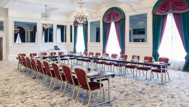 Konferenssihuone, jossa on pitkä pöytä, punaiset tuolit, koristeelliset kattokruunut ja suuret ikkunat, joissa on verhot.