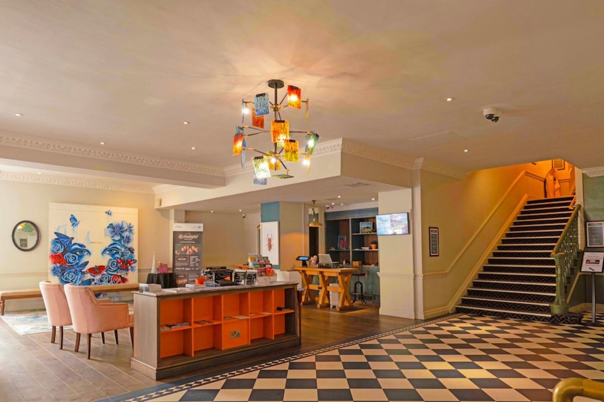A hotel lobby with a checkered floor, a reception desk, a staircase, and a colorful ceiling light fixture.