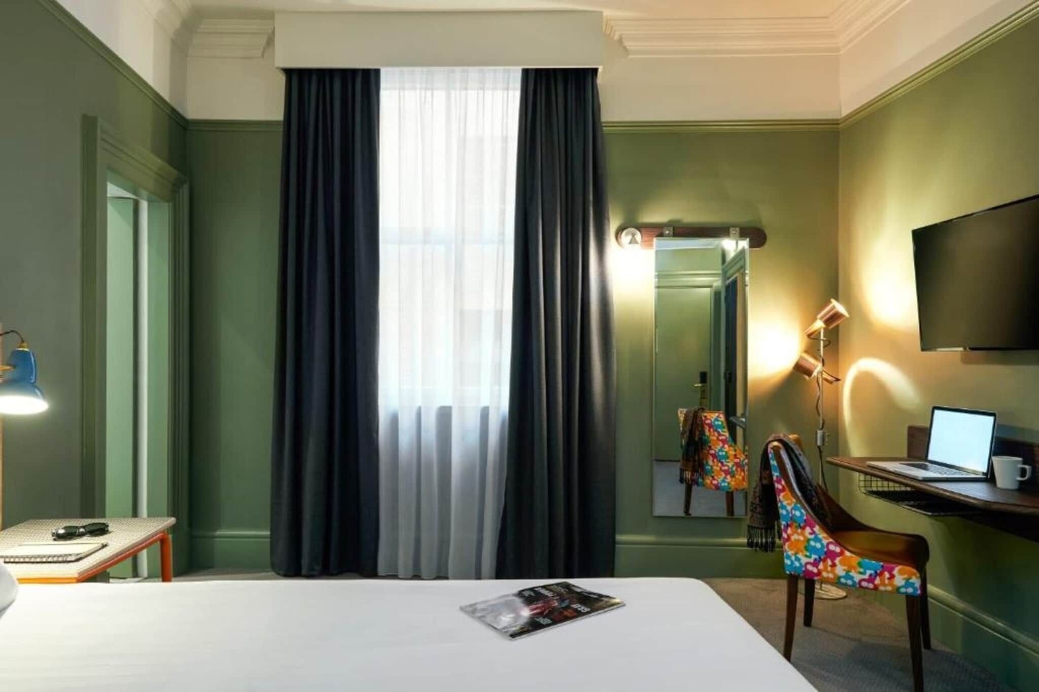 A hotel room with a bed, a desk with a laptop, a chair, a lamp, and a television on the wall.