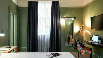 A hotel room with a bed, a desk with a laptop, a chair, a lamp, and a television on the wall.