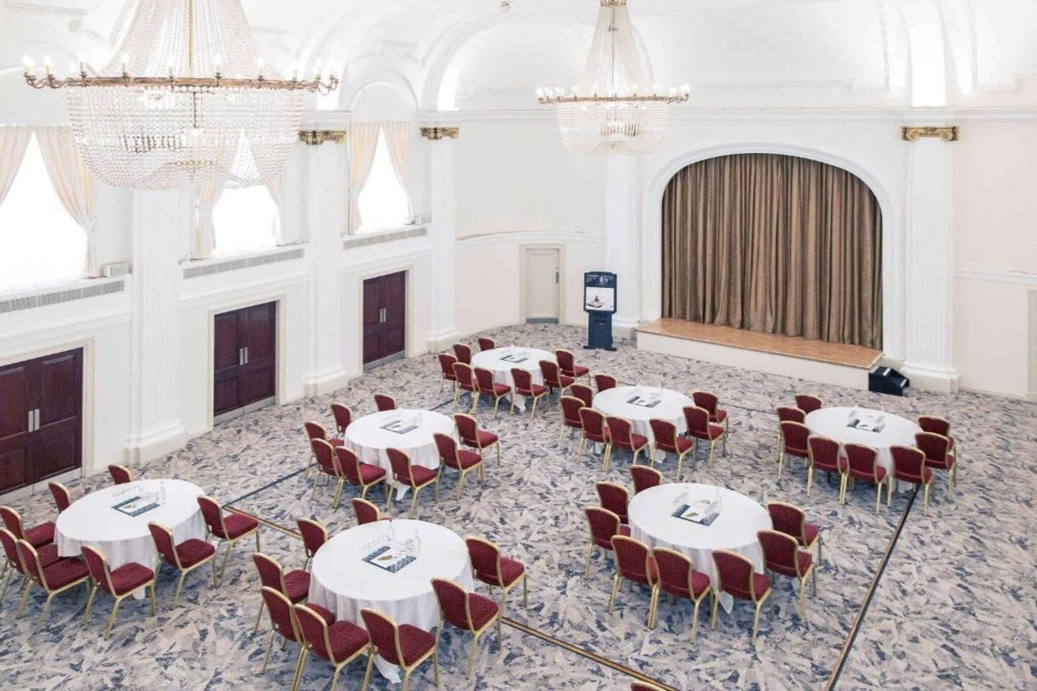 A conference room with round tables and red chairs, chandeliers, and a stage with curtains.