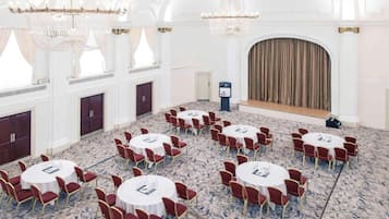 A conference room with round tables and red chairs, chandeliers, and a stage with curtains.