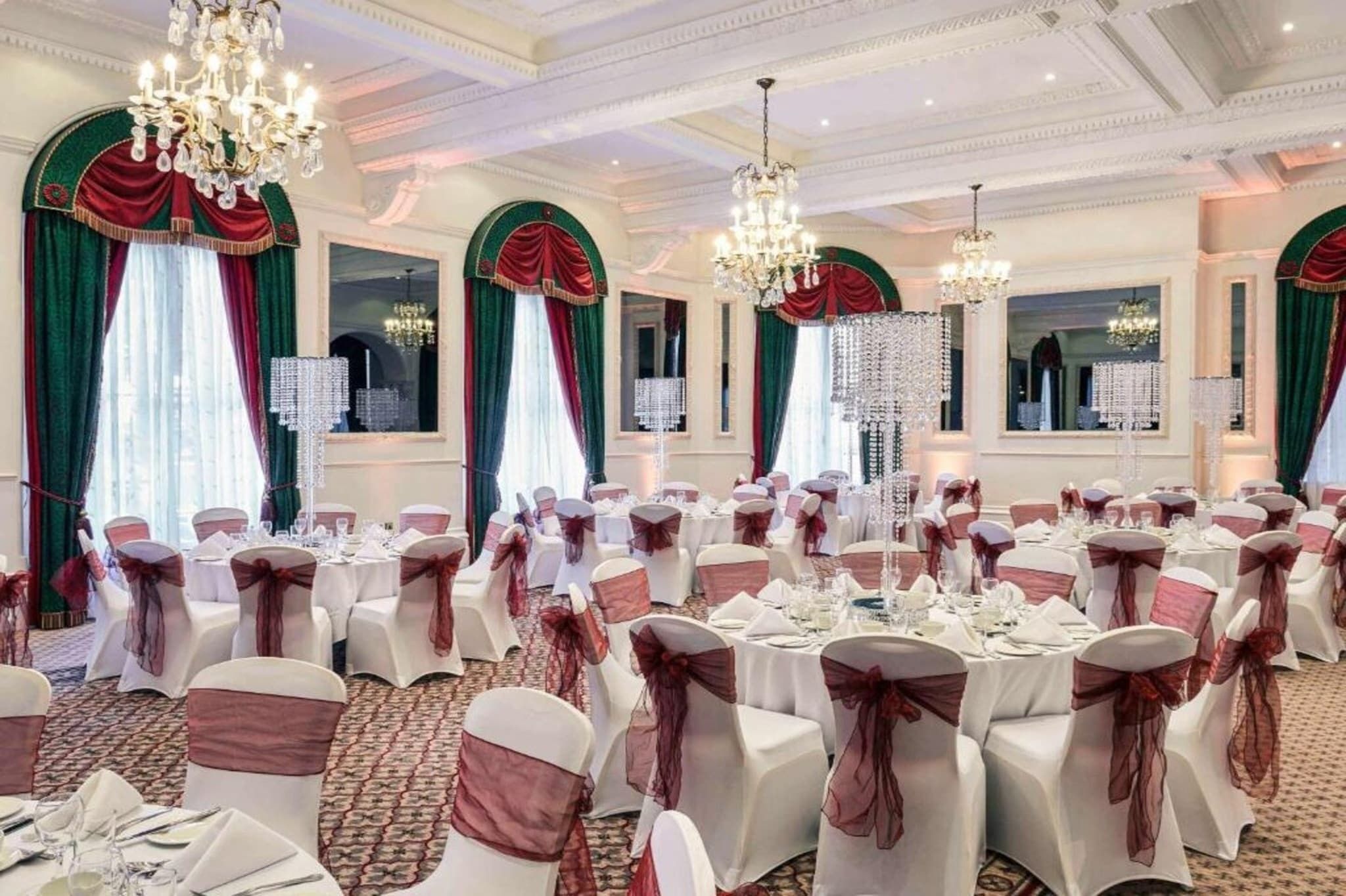 A banquet hall with round tables, chairs with sashes, chandeliers, and draped windows.