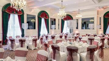 A banquet hall with round tables, chairs with sashes, chandeliers, and draped windows.
