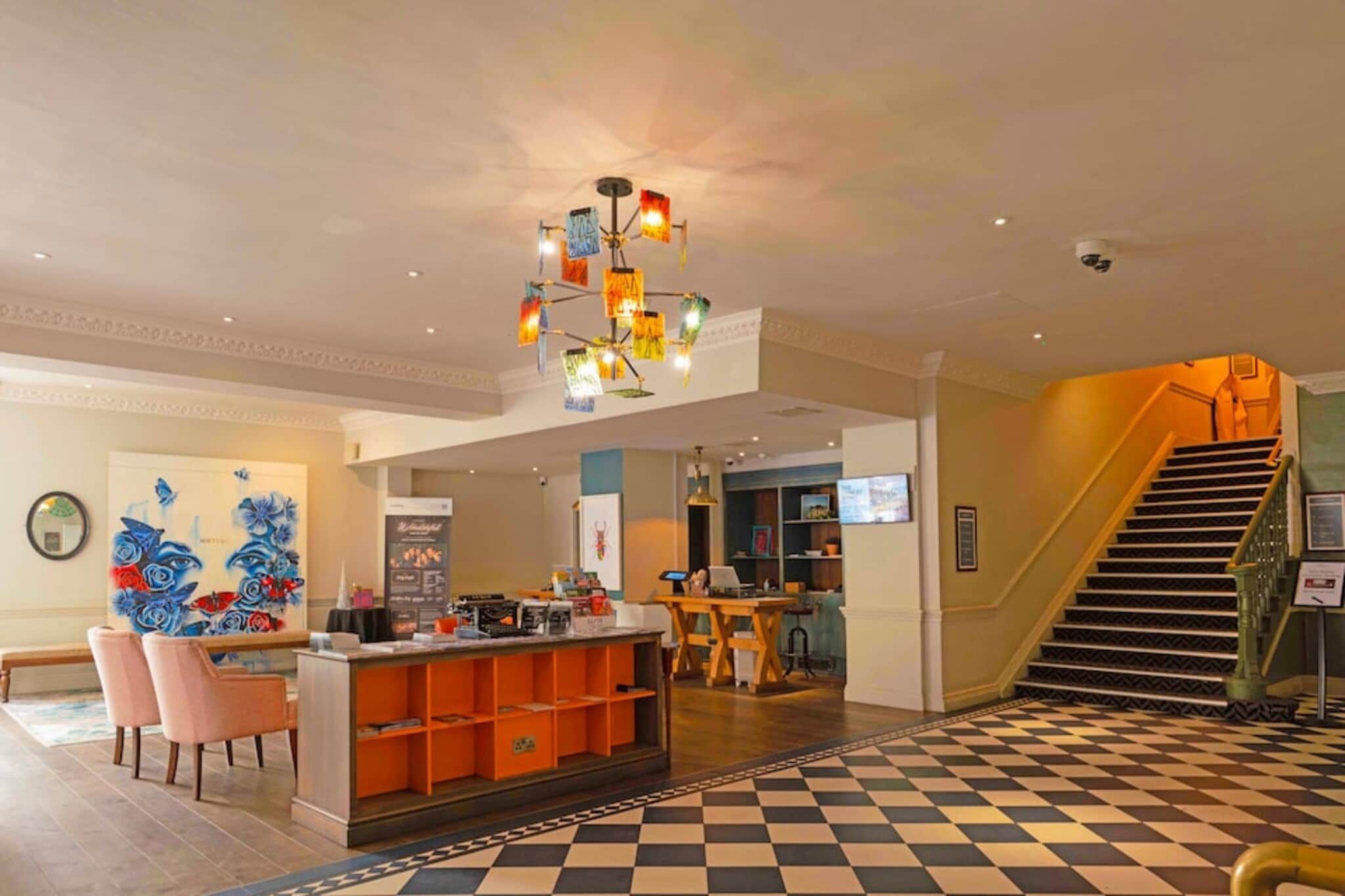 A hotel lobby with a checkered floor, a reception desk, a staircase, and a wall with a floral mural.
