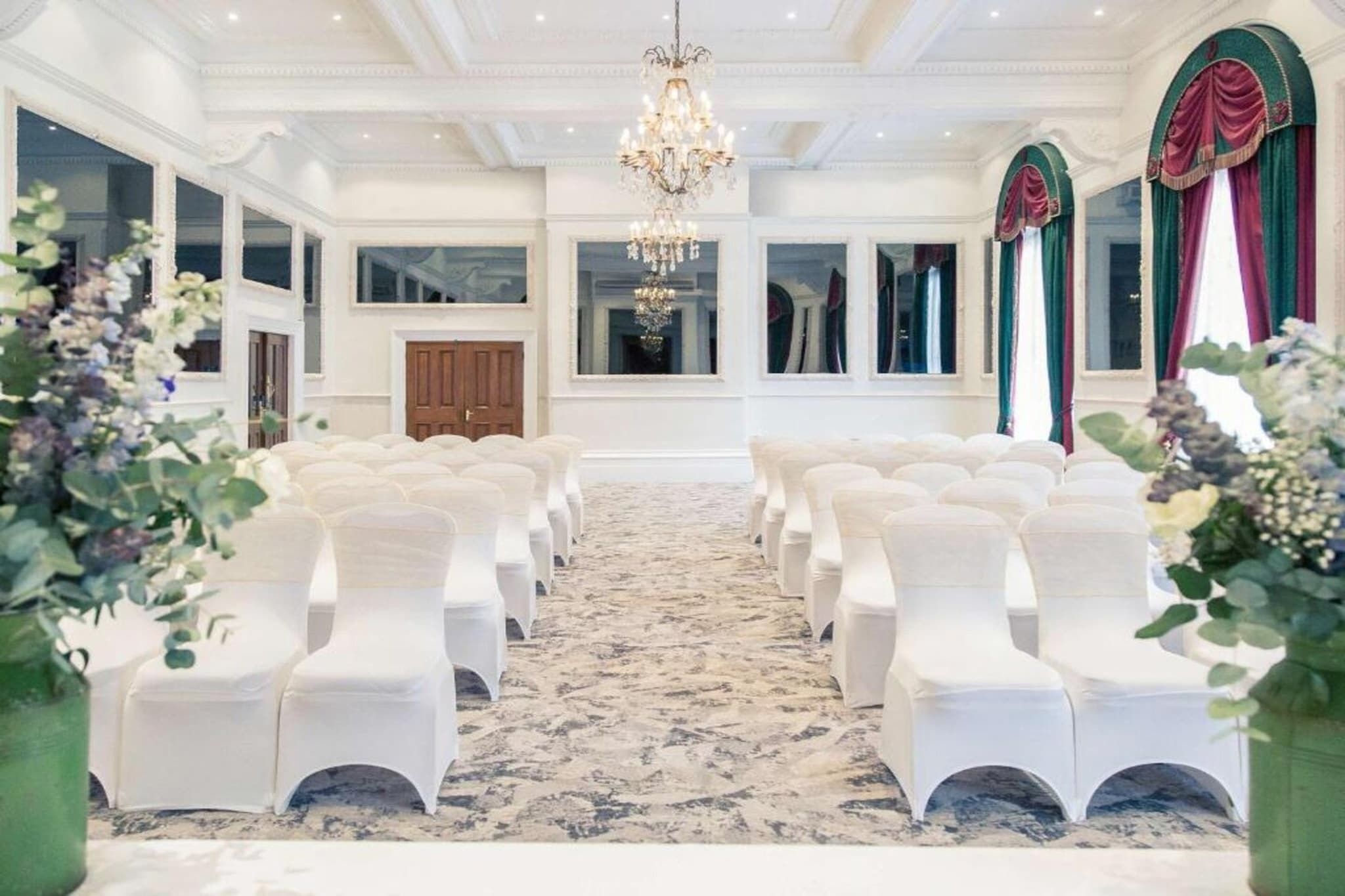 A formal indoor setting with rows of white chairs, a chandelier, and large windows with curtains.