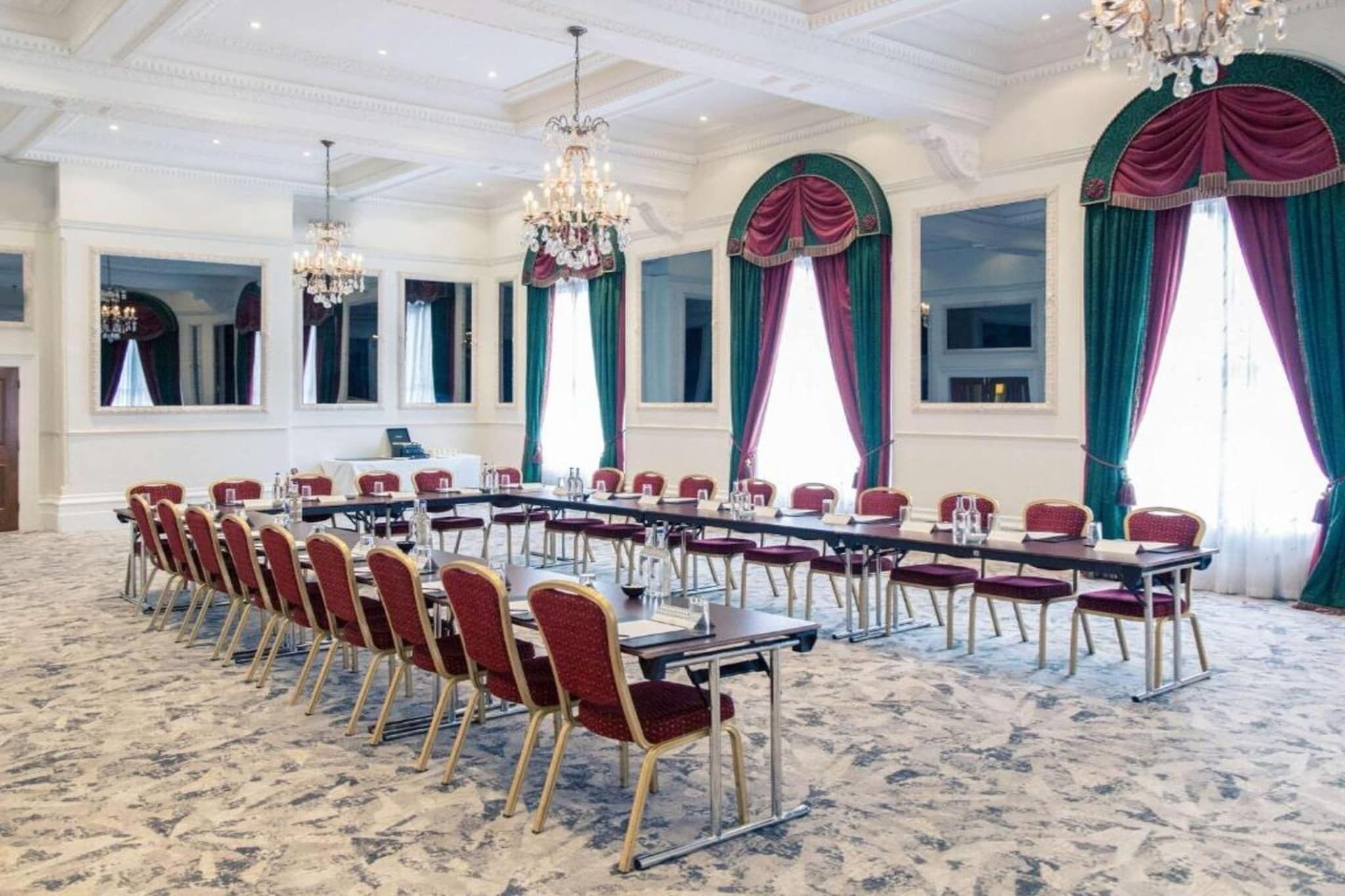 A conference room with a long table, red chairs, chandeliers, and large windows with curtains.