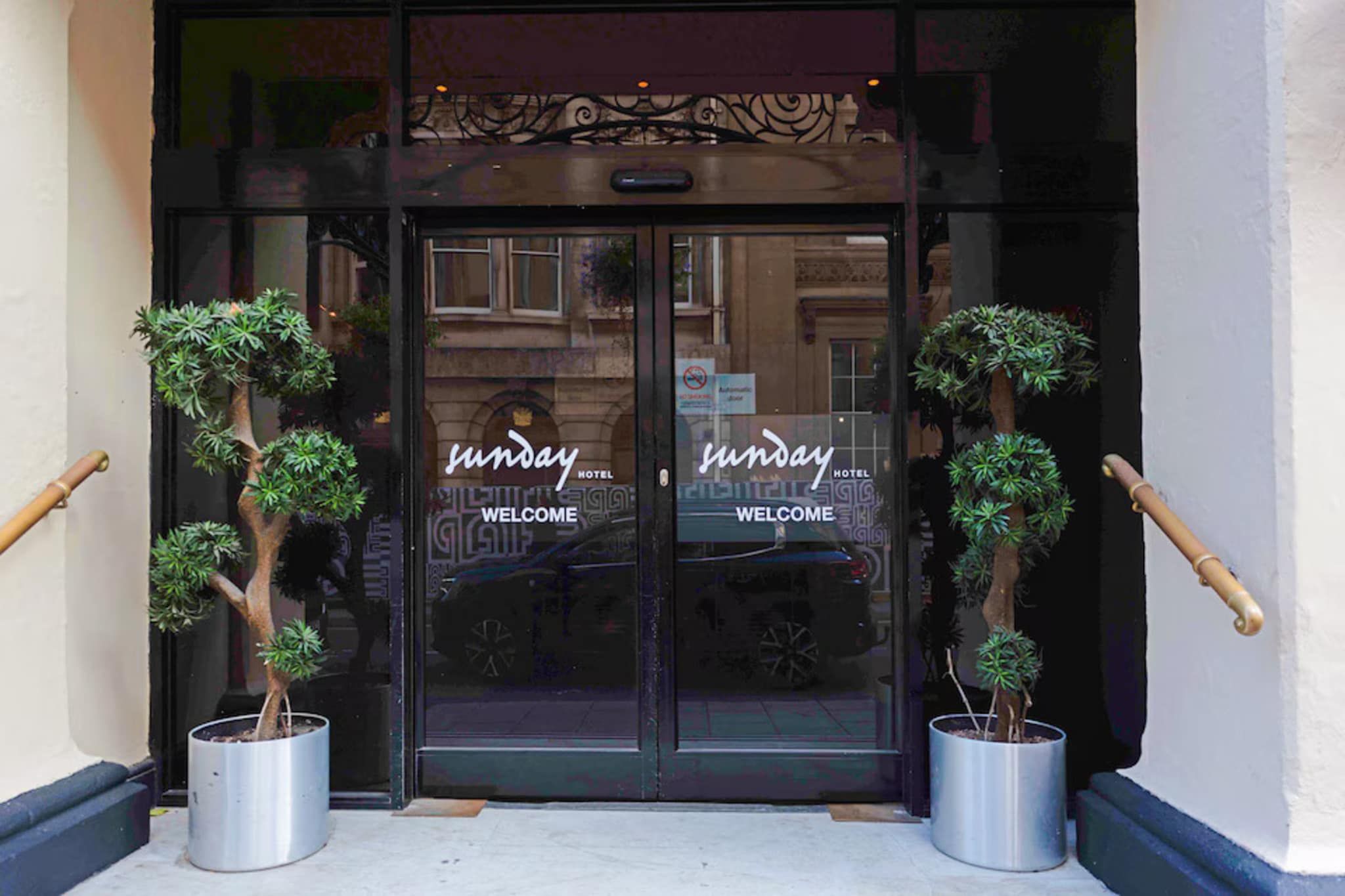 A hotel entrance with a glass door, two potted plants, and the name 'Sunday Hotel' on the glass.