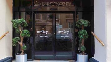 A hotel entrance with a glass door, two potted plants, and the name 'Sunday Hotel' on the glass.