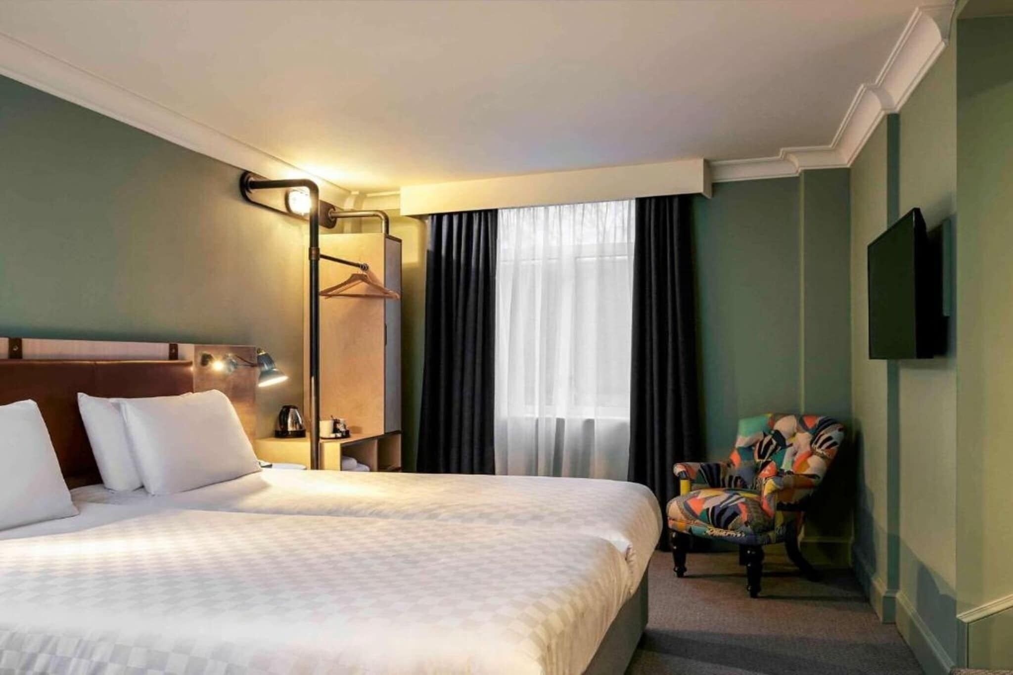 A hotel room with a bed, a chair, a coat rack, a television, and a window with curtains.