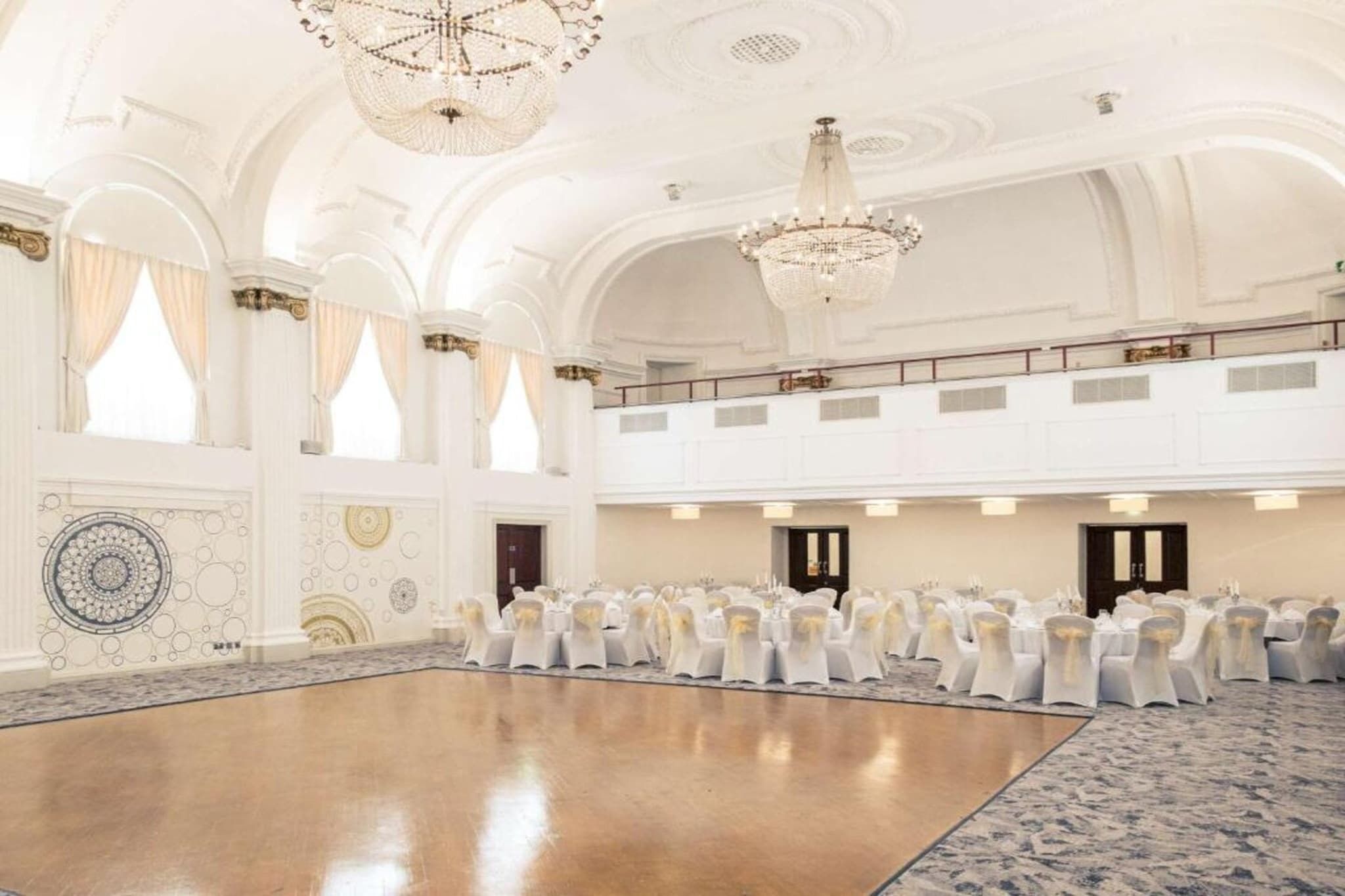 A spacious hall with a polished floor, white chairs with gold sashes, and ornate chandeliers.
