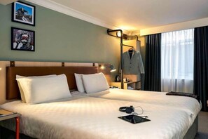 Una habitació d'hotel amb un llit gran, una taula de nit, una làmpada, un penjador i una finestra amb cortines.