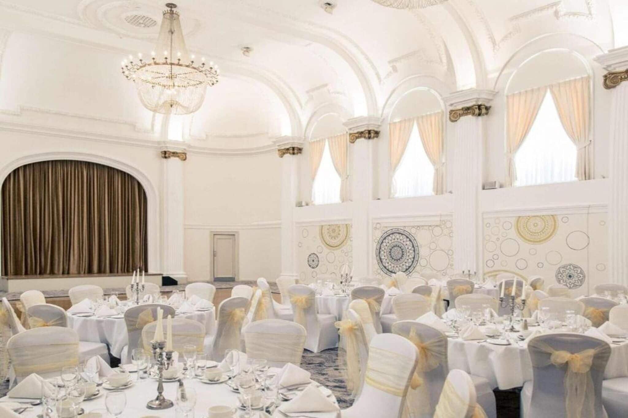 A banquet hall with round tables, white chairs, chandeliers, and decorative wall patterns.