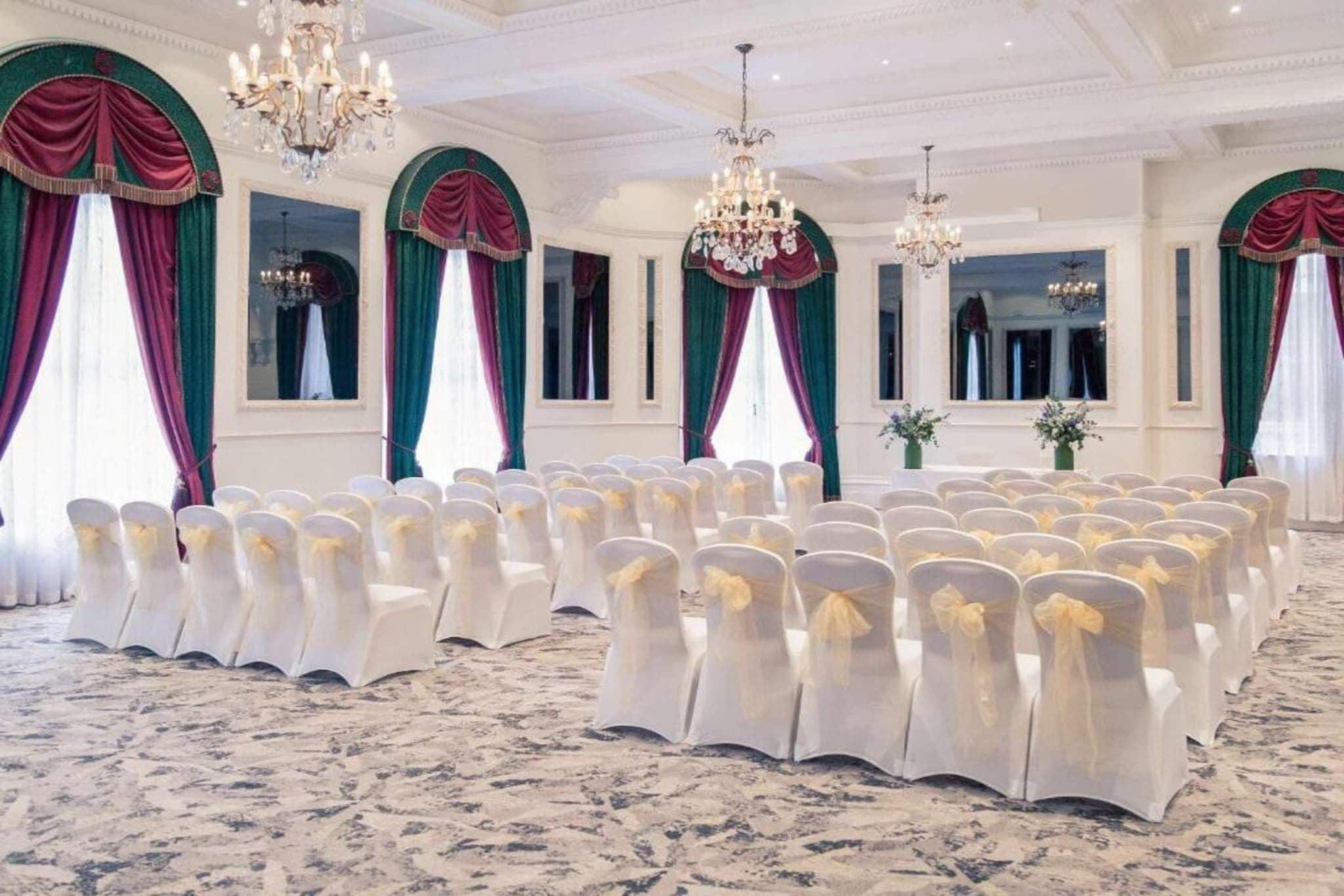 A formal event space with rows of chairs, chandeliers, and draped windows.