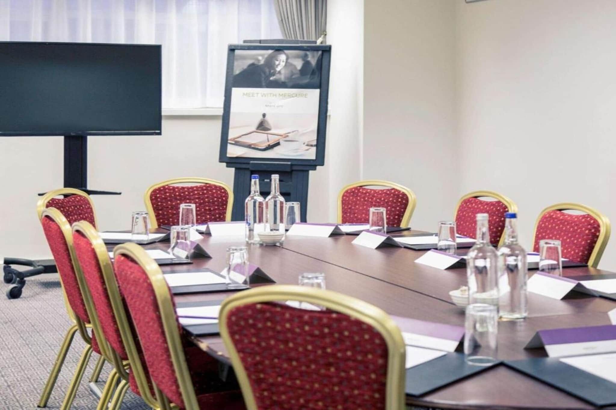 A conference room with a long table, red chairs, water bottles, and a projector screen.