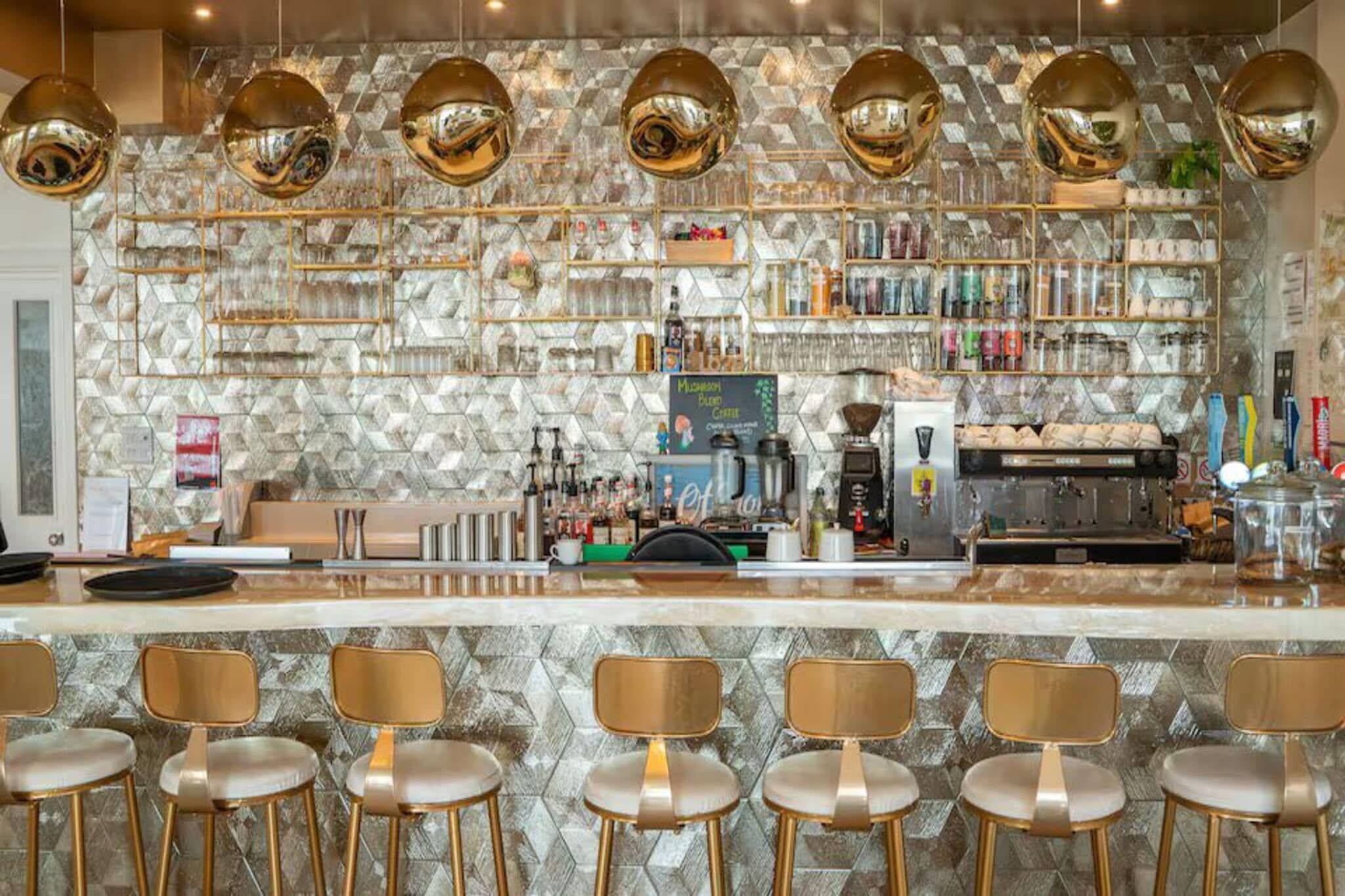 Un espace bar avec un mur décoré d’un motif en pierre, plusieurs luminaires suspendus, une machine à café et des tabourets de bar.