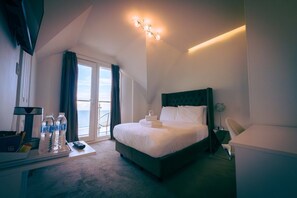 A hotel room with a bed, a desk with a water bottle, a remote control, and a view of the sea through the window. - Newquay Beach by Sunday - Compact Single Room (Newquay)