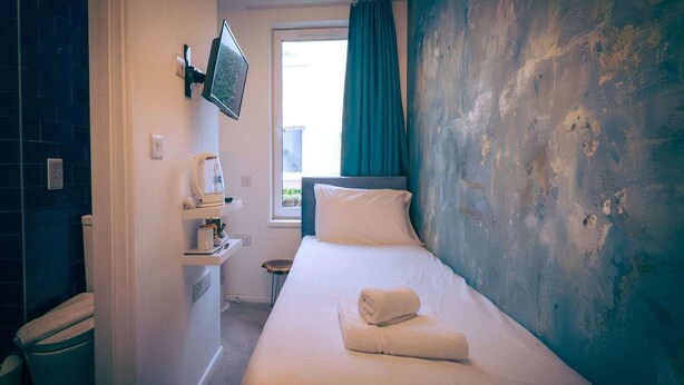 Habitación de hotel con una cama, un televisor montado en la pared, una ventana con cortinas y una mesita con hervidor.
