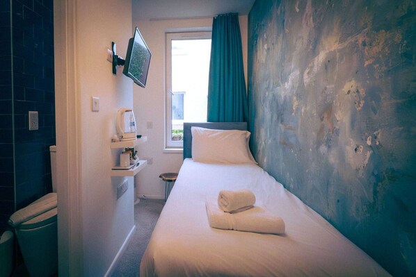 A hotel room with a bed, a television mounted on the wall, a window with curtains, and a small table with a kettle. - Standard Single Room (Newquay)