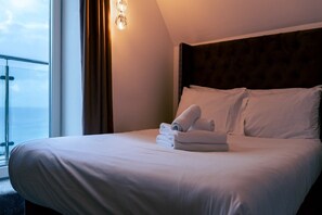A neatly made bed with pillows and a folded towel, a window with curtains, and a wall-mounted light fixture. - Executive Double with Balcony & Sea View (Newquay)