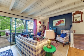 Ein gemütliches Wohnzimmer mit einem karierten Sofa, einem gestreiften Sessel, einem grünen Beistelltisch und einem großen Fenster mit Blick auf Grünflächen.