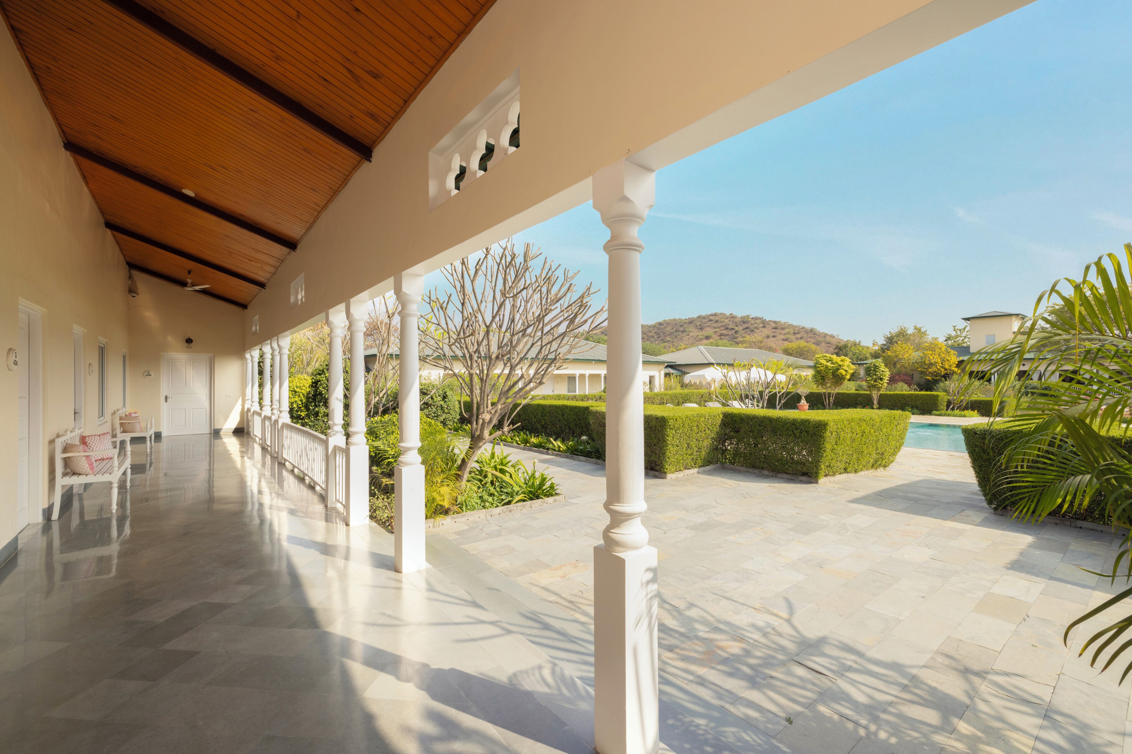 A covered outdoor area with a wooden ceiling, white columns, and a view of a landscaped garden with a pool.
