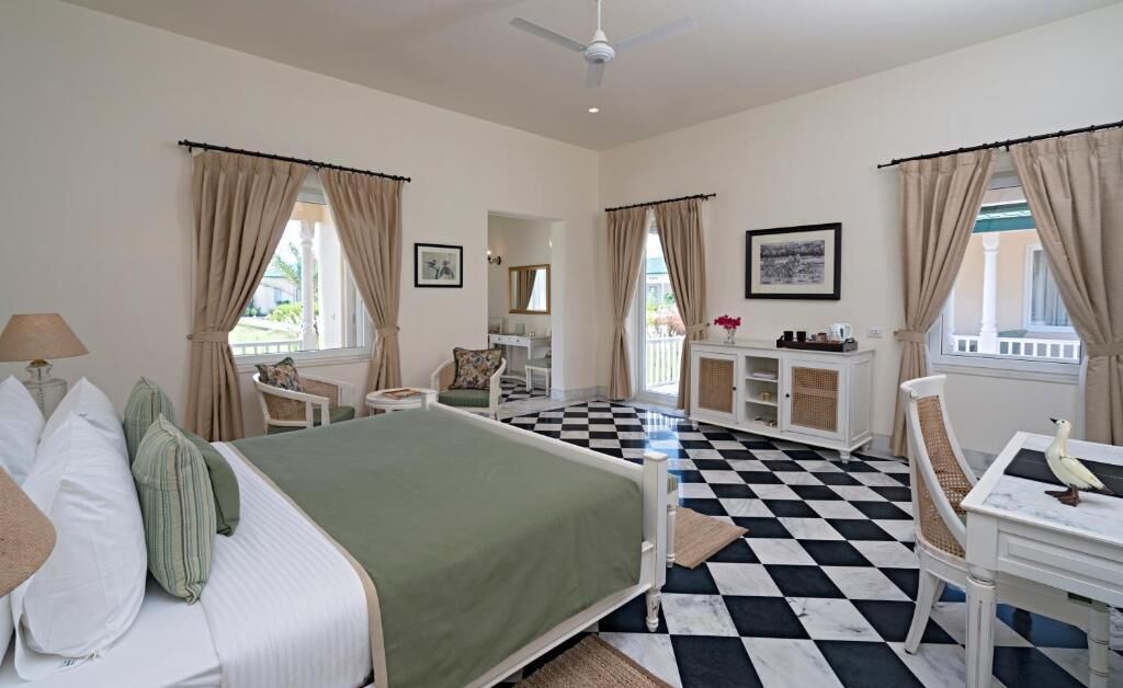 A hotel room with a bed, a desk, a chair, a window with curtains, a ceiling fan, and a checkered floor.