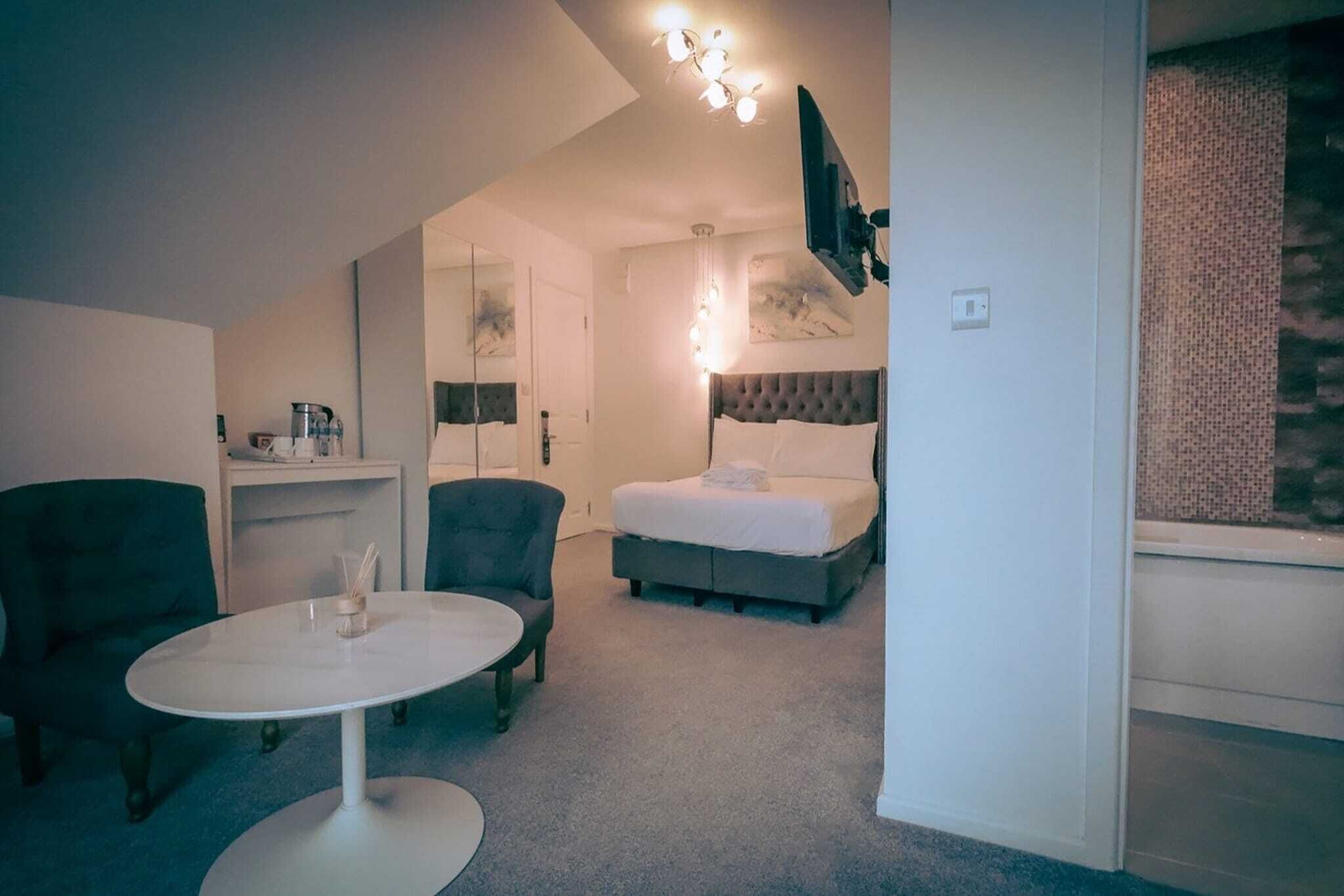 Habitación de hotel con cama, dos sillones, una mesa redonda, un espejo, televisor montado en la pared y una kitchenette pequeña.