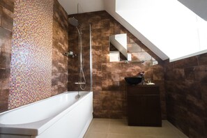 Bany modern amb paret de rajola mosaic, banyera independent, recinte de dutxa de vidre i un moble de tocador de fusta fosca.