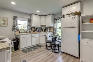 A kitchen with white cabinets, a refrigerator, a dining table with chairs, and a window with curtains.