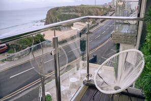 Un balcó amb una cadira blanca, vista d'una carretera costanera i un indici de l'oceà.