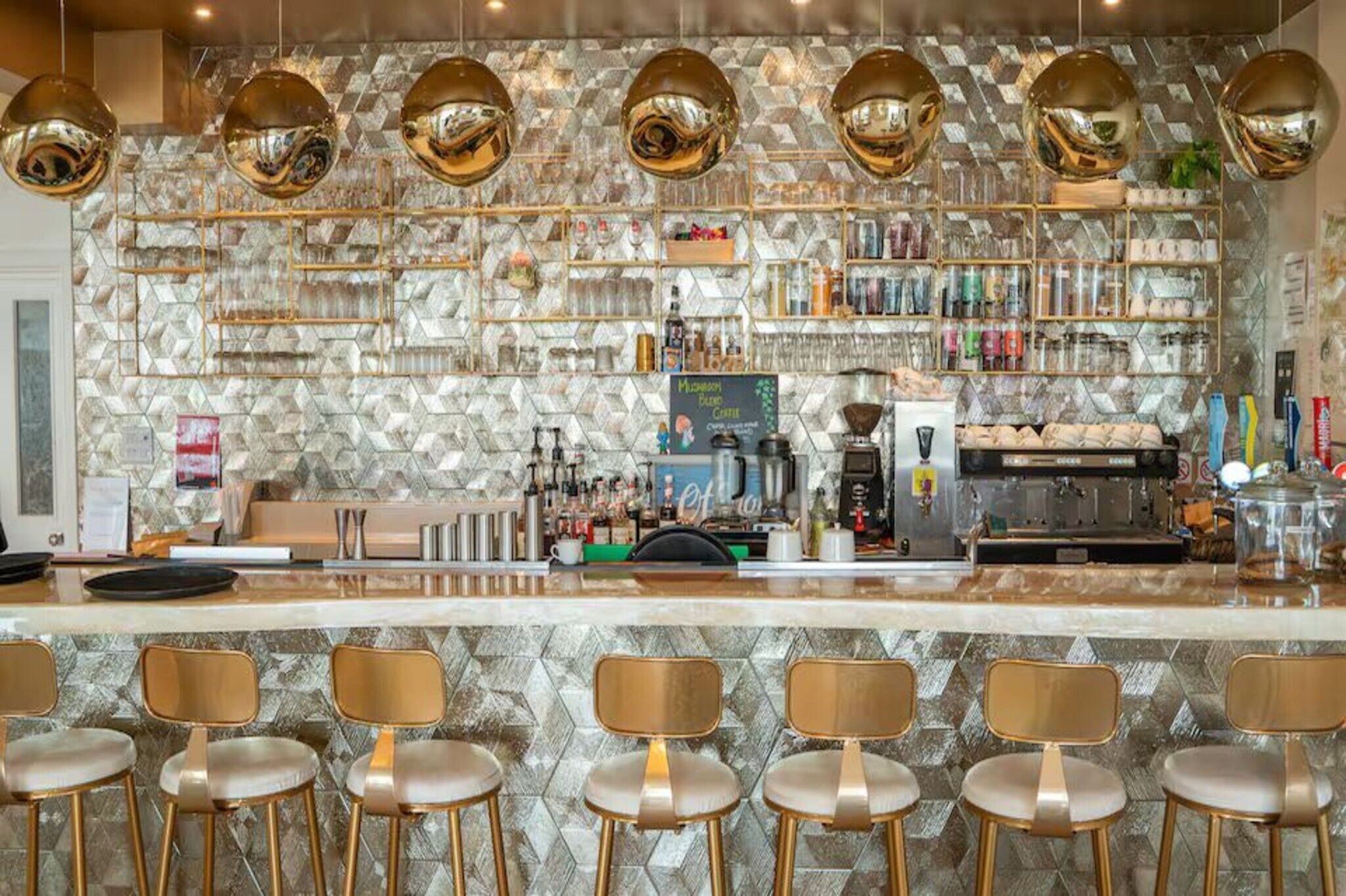 Eine Bar mit einer Steinwand, Regalen voller Flaschen, einer Kaffeemaschine und goldenen Stühlen.