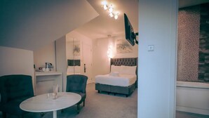 Quarto de hotel com cama, duas poltronas, uma mesa redonda, espelho, televisão na parede e uma pequena cozinha equipada.