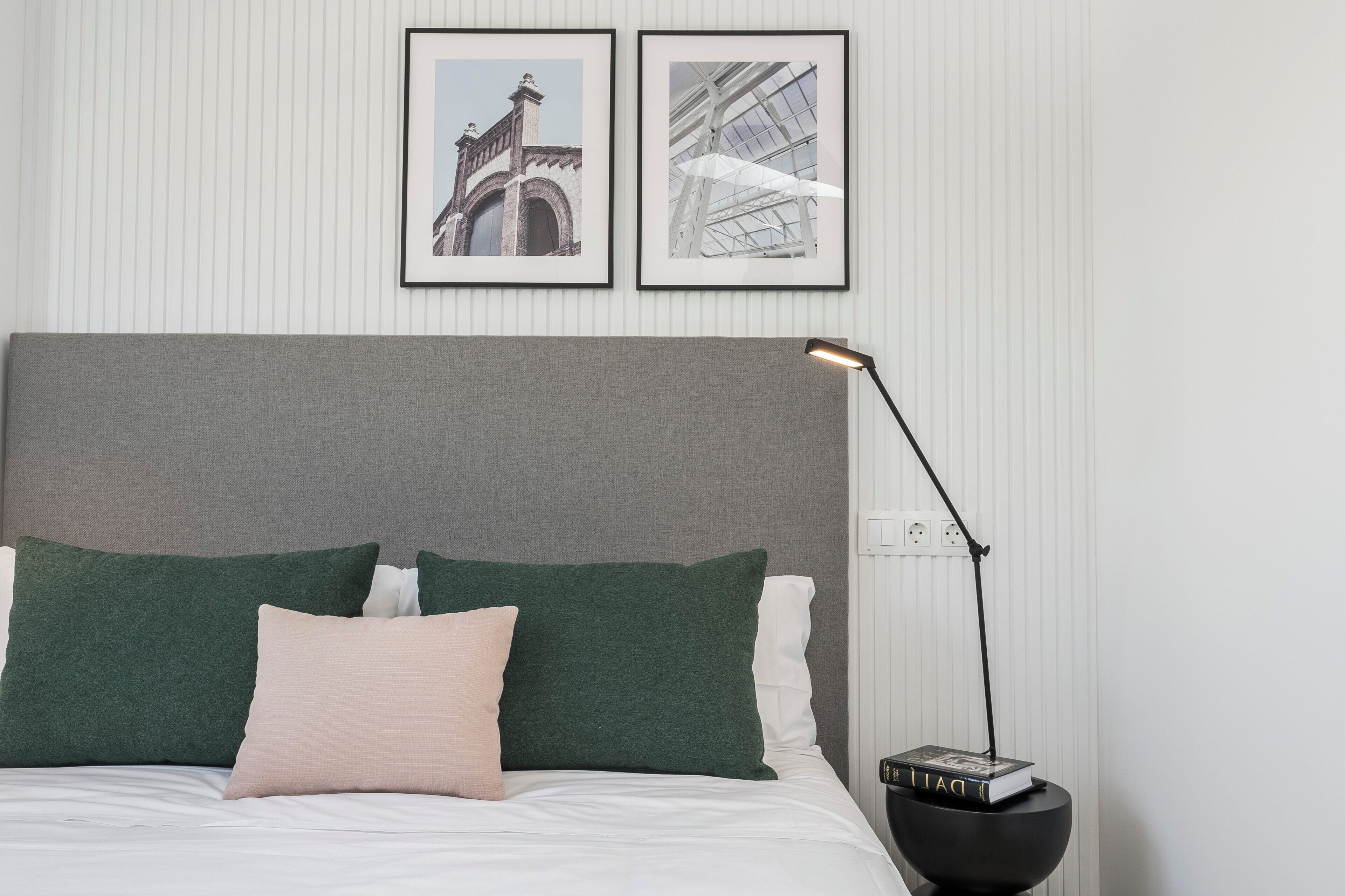 Una cama bien hecha con cabecera gris, almohadas verdes y blancas y una lámpara de noche negra.