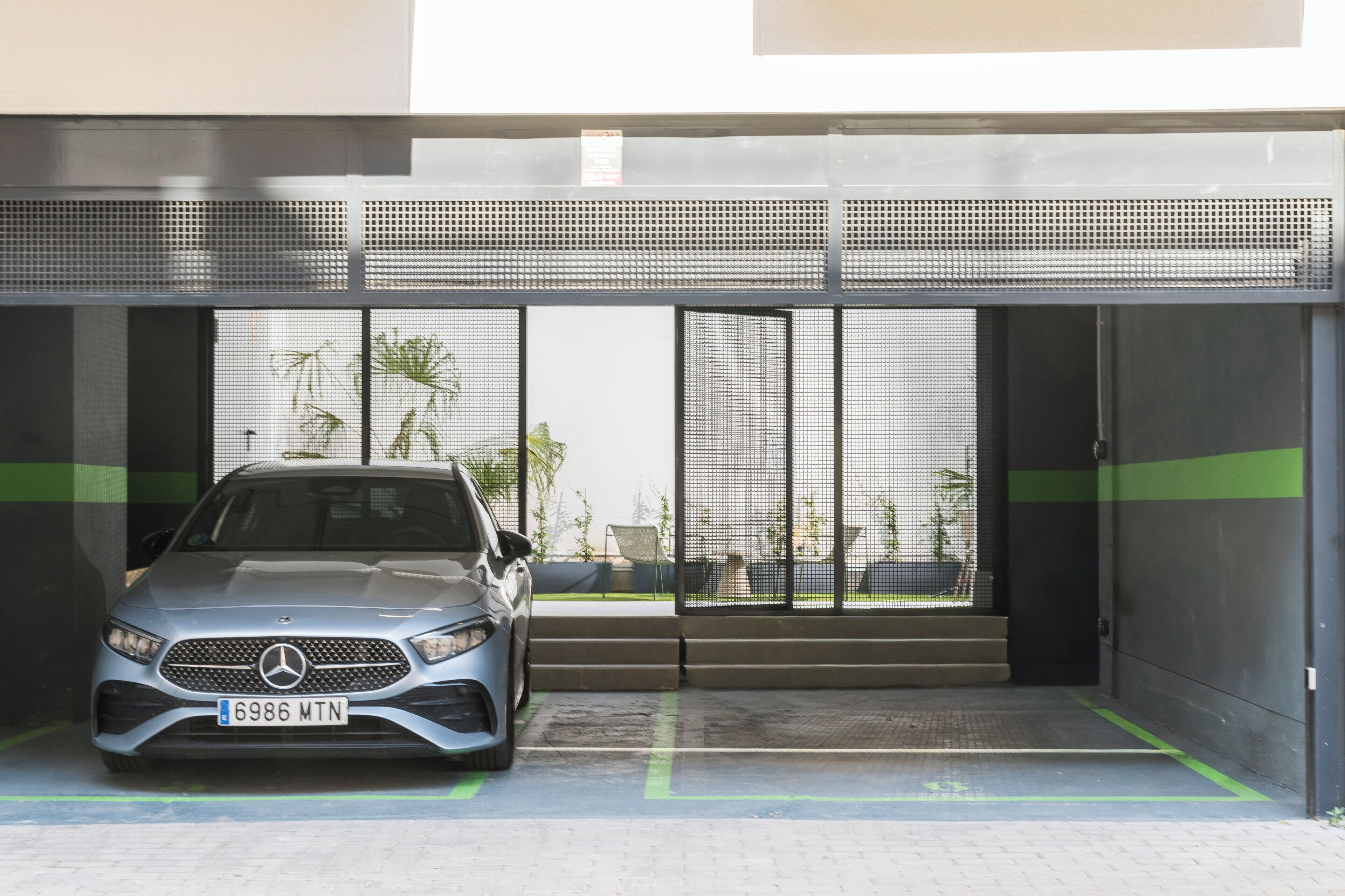 Un coche estacionado en un área de estacionamiento techada con diseño moderno, que incluye una puerta metálica y una franja verde en el suelo.