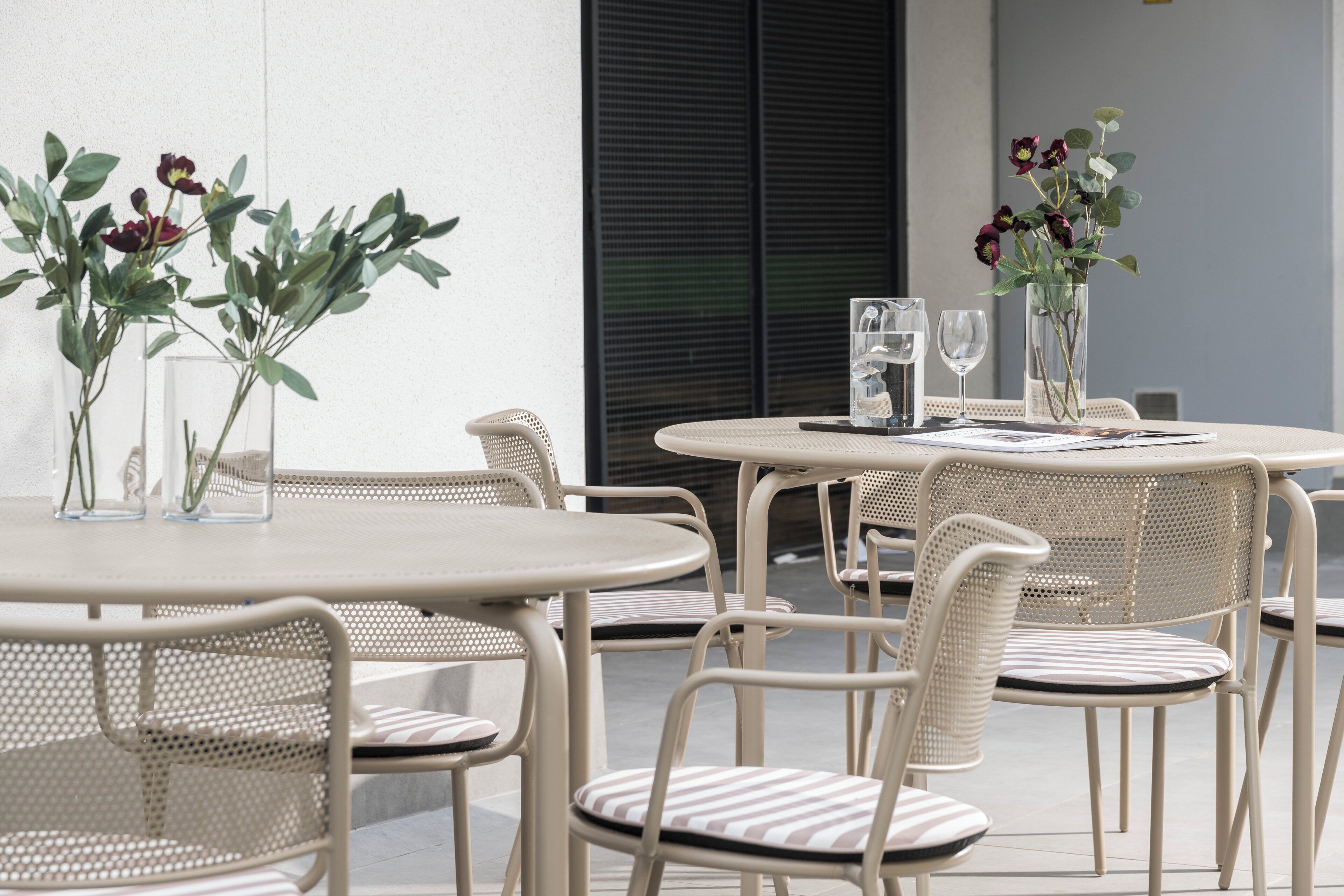 Een terras met ronde tafels en stoelen, een glazen vaas met bloemen en een glas water op de tafel.