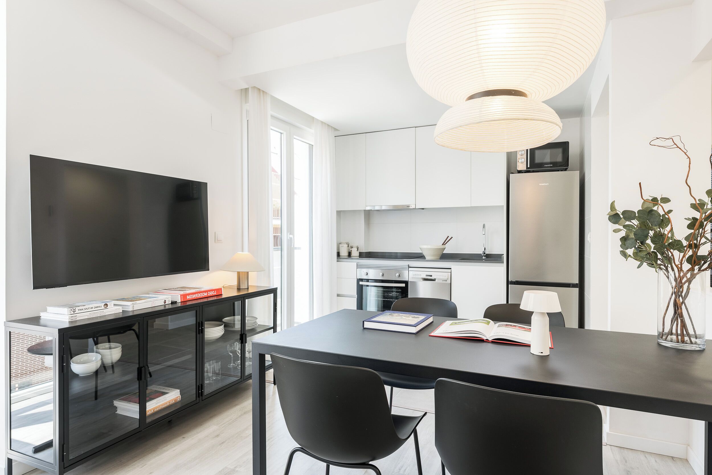 A modern kitchen with a flat-screen TV, dining table, chairs, and a pendant light.