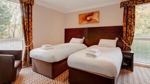 A hotel room with two beds, a painting on the wall, and a view of trees through the window.
