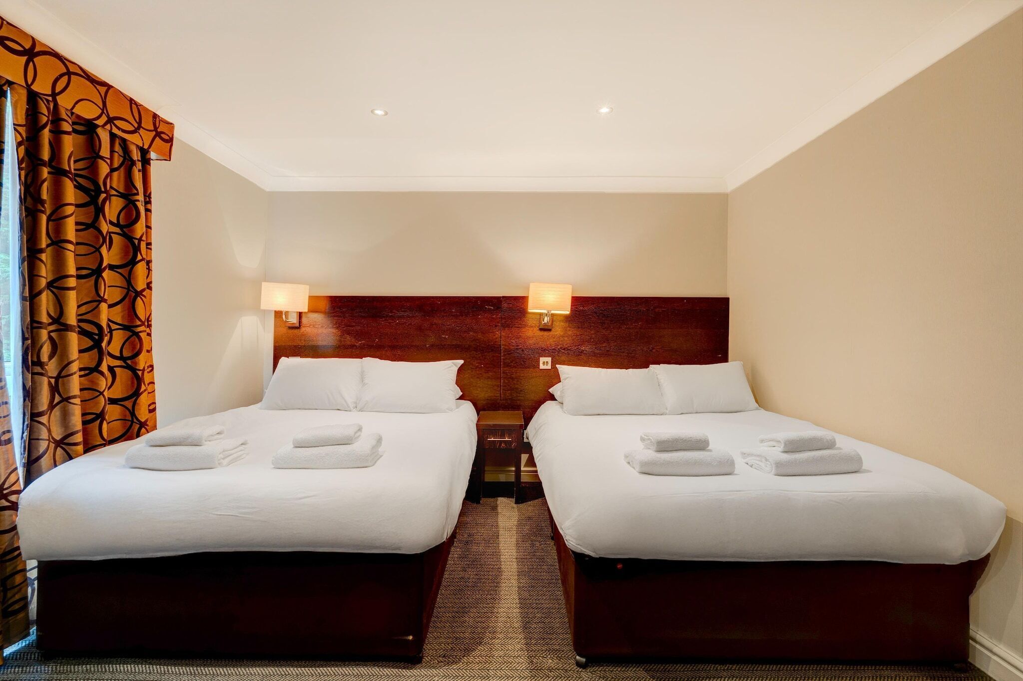 A hotel room with two beds, each with white linens and towels, a wooden headboard, and two wall-mounted lamps.