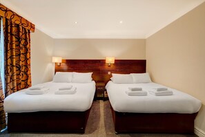 Quarto de hotel com duas camas, cada uma com roupa de cama e toalhas brancas, uma cabeceira de madeira e duas luminárias fixadas na parede.