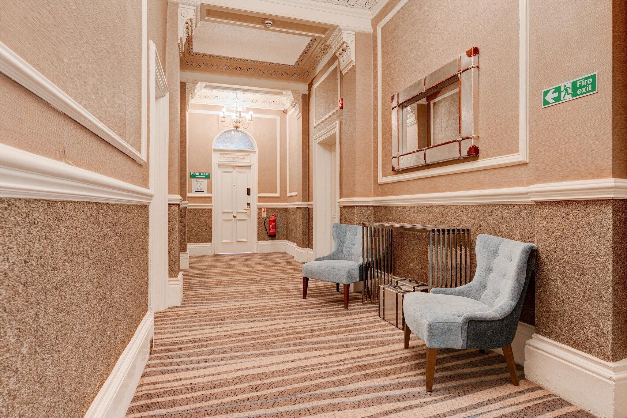 Um corredor de hotel com piso carpeteado, dois poltronas, uma placa de saída de incêndio e uma porta com a inscrição 'The Director's Suite'.