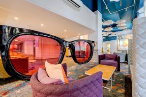 A lobby with large, black-framed mirrors, a seating area with purple and orange cushions, and a ceiling with hanging umbrellas.