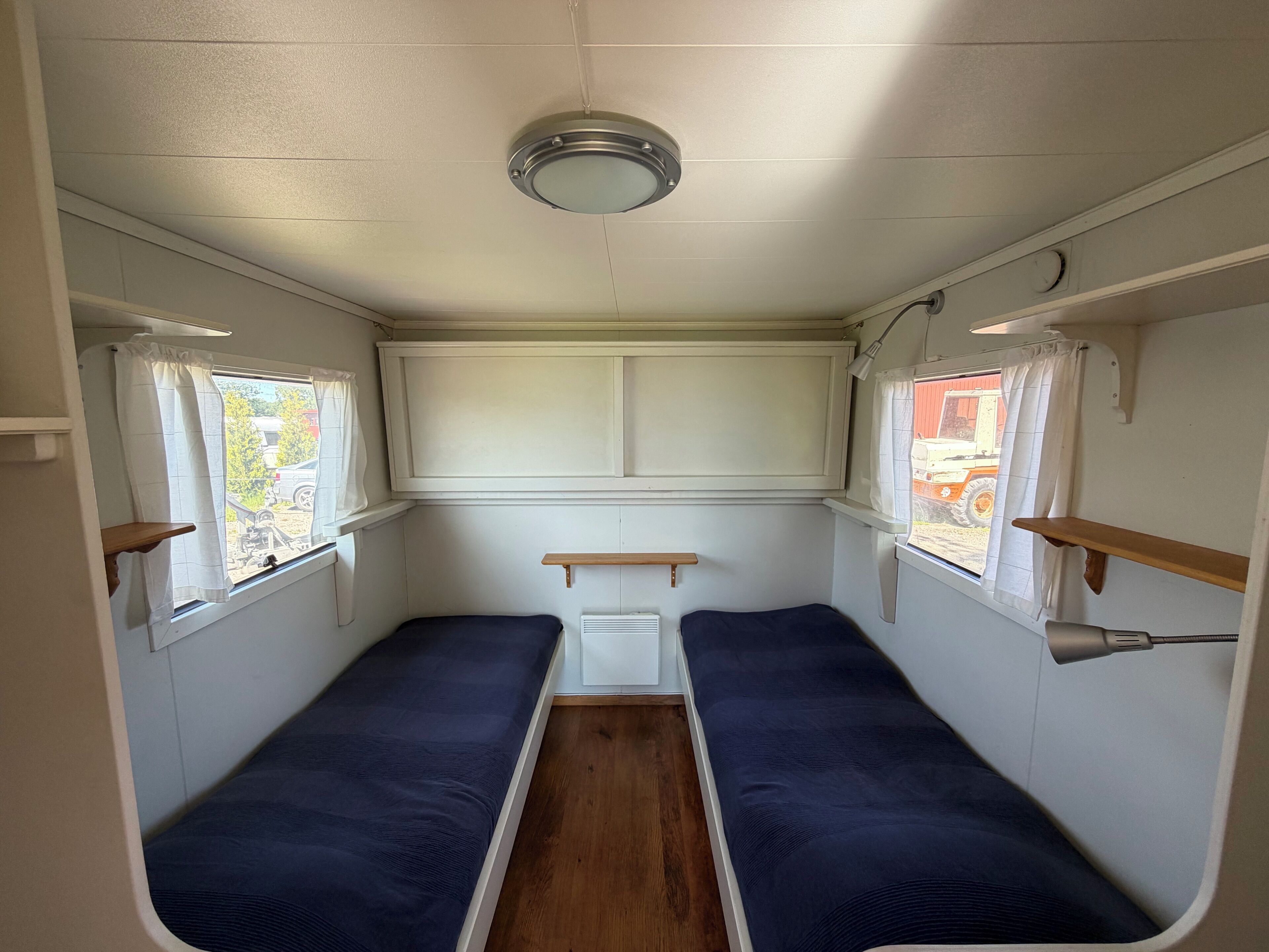 A narrow interior with two single beds, a window with curtains, a white ceiling light, and a small white cabinet.