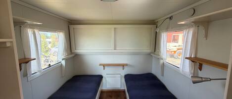 A narrow interior with two single beds, a window with curtains, a white ceiling light, and a small white cabinet.