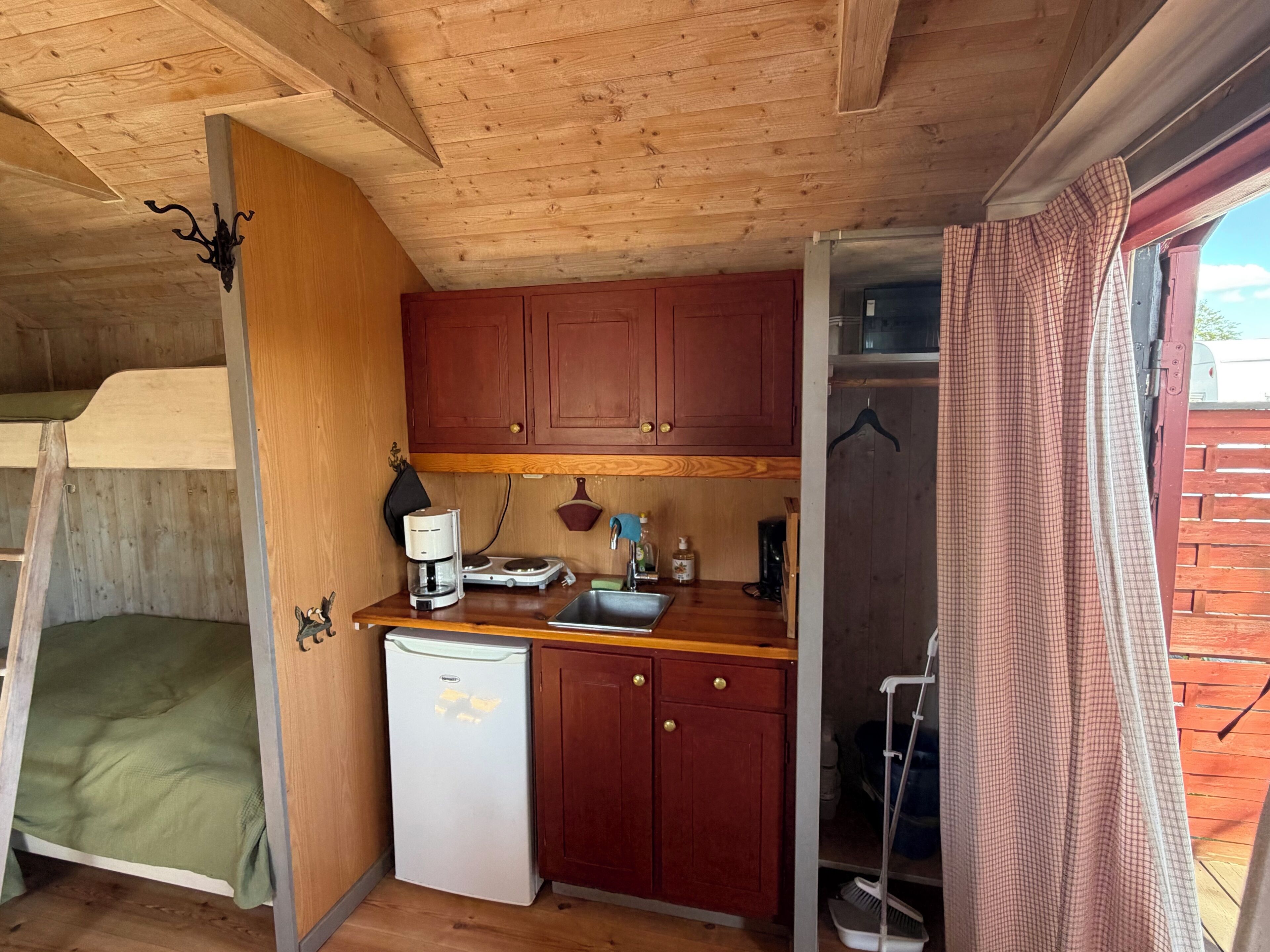 A compact kitchen with wooden cabinets, a small refrigerator, a coffee maker, and a broom.