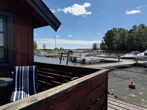 Una terraza de madera con un cojín a rayas, vista a un puerto deportivo con barcos amarrados y un cielo despejado.