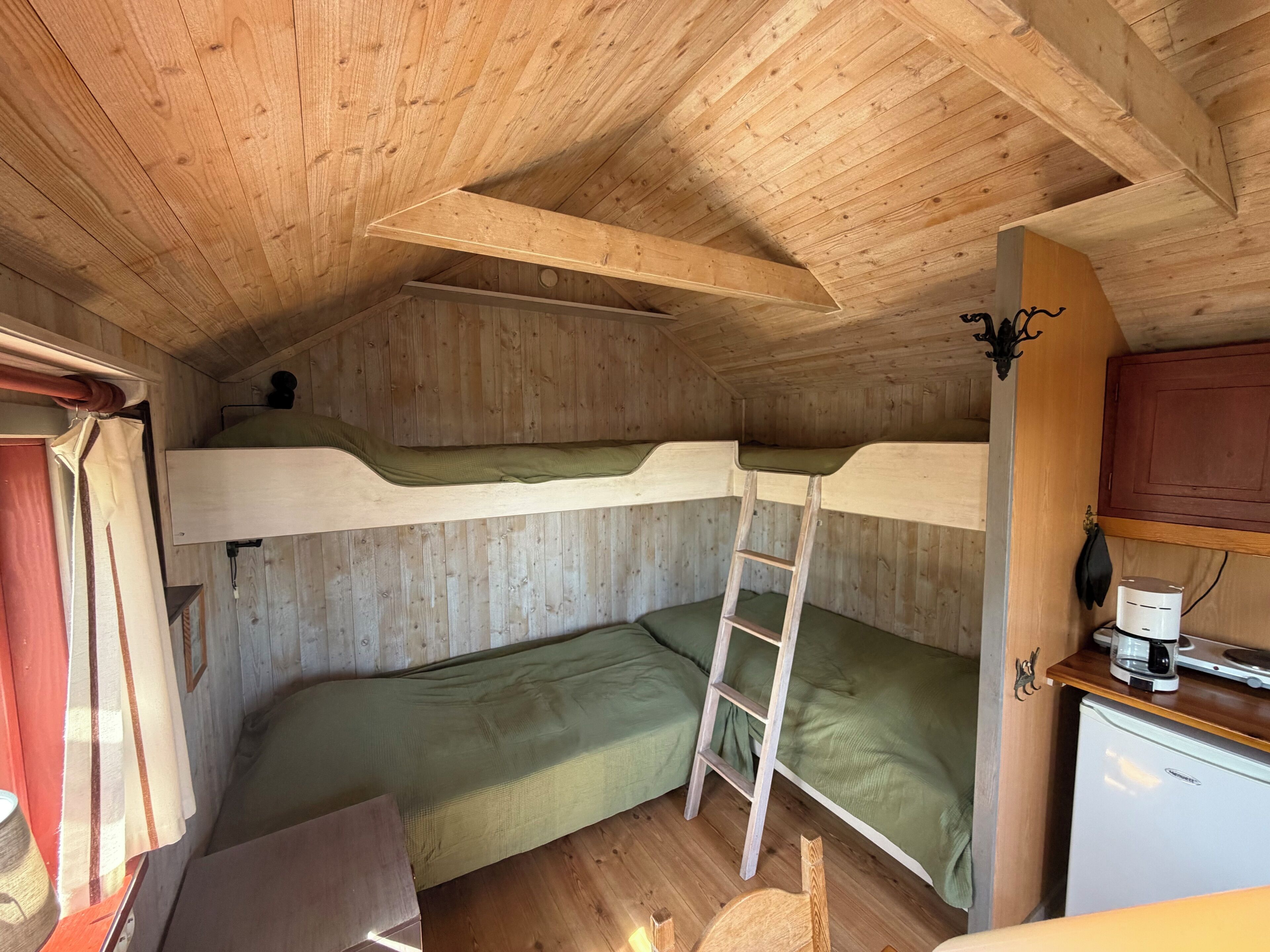 Interior de cabana com paredes de madeira, beliche, escrivaninha, escada e uma pequena geladeira.
