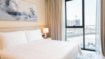 Habitación de hotel con una cama grande, una mesita de noche con lámpara, ventana con vista a la ciudad y un cuadro en la pared.