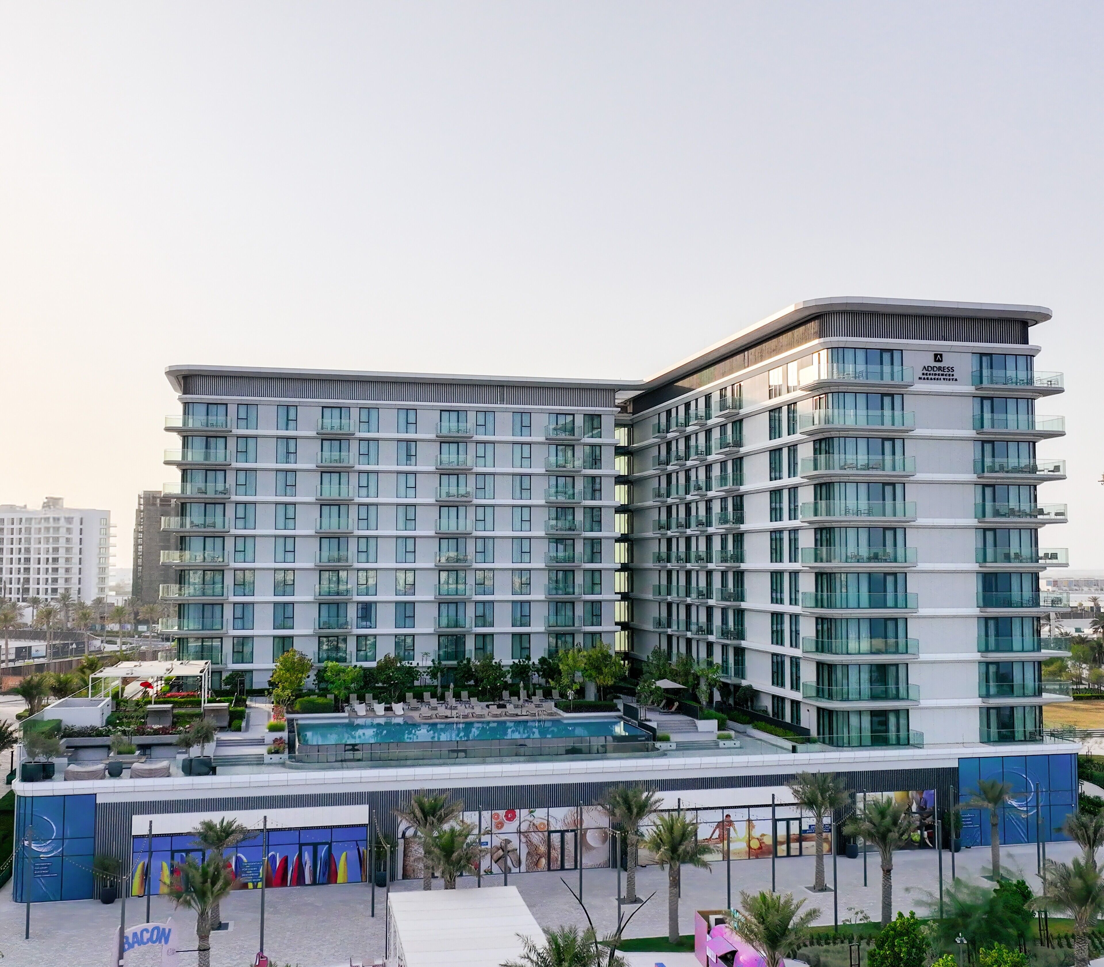 Ein modernes Hotel mit mehreren Etagen, einem Schwimmbad und einer angelegten Grünfläche mit Palmen.